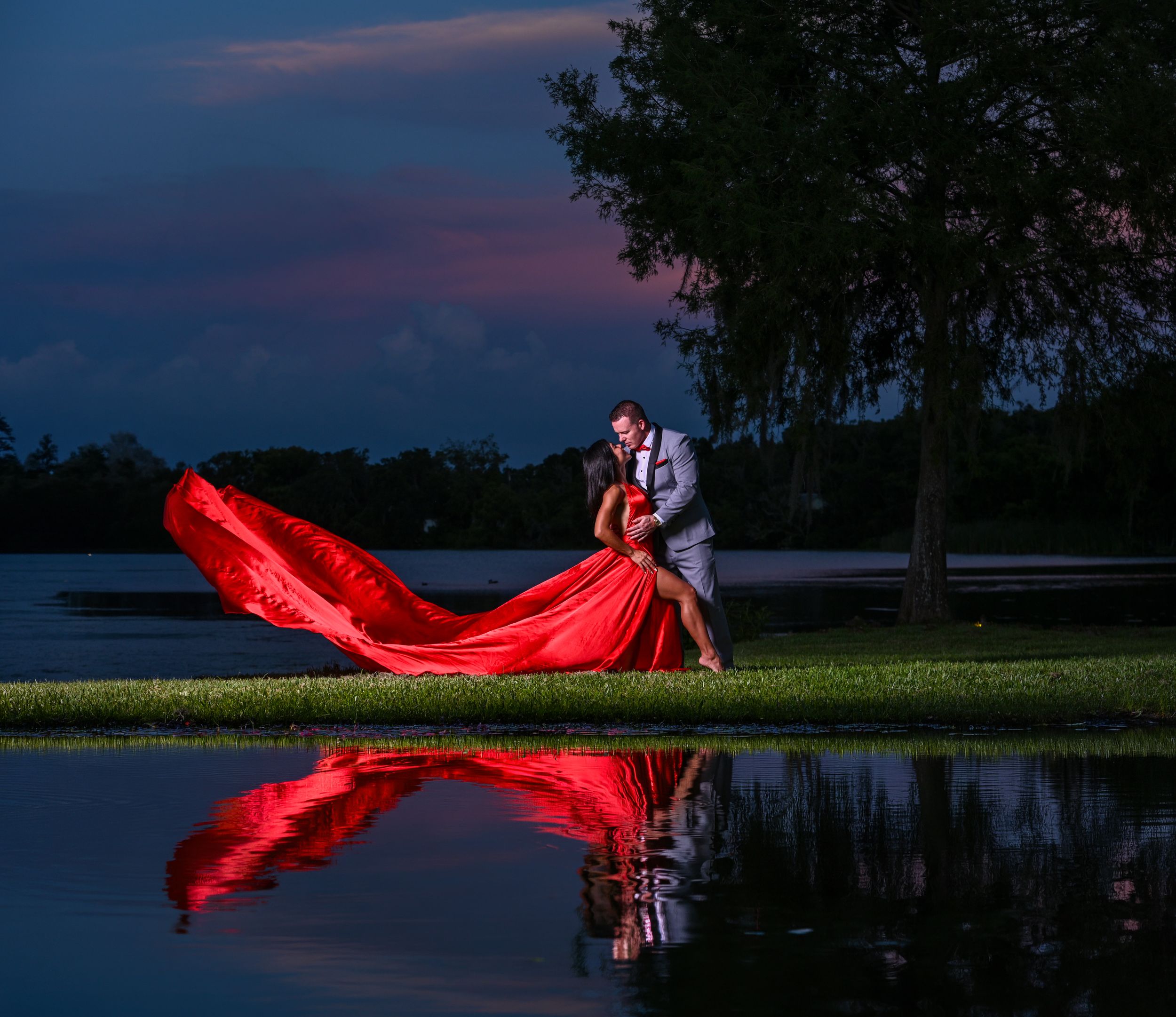 Couple dancing at dusk with woman in dramatic red gown reflected in still water near lake.