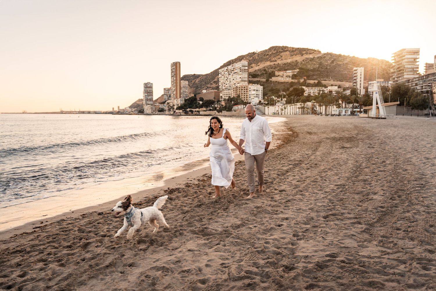 Couple smiling and laughing and chasing their dog on the beach at a sunset photo shoot in Alicante Spain
