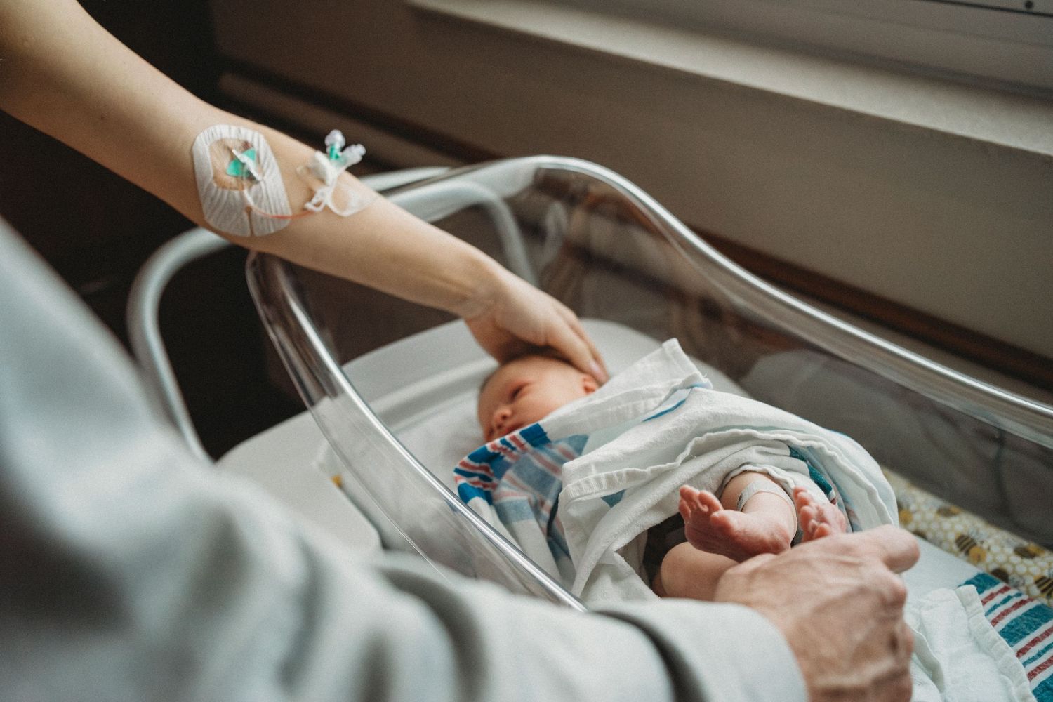Hands tend to newborn baby connected to medical equipment in hospital bed.