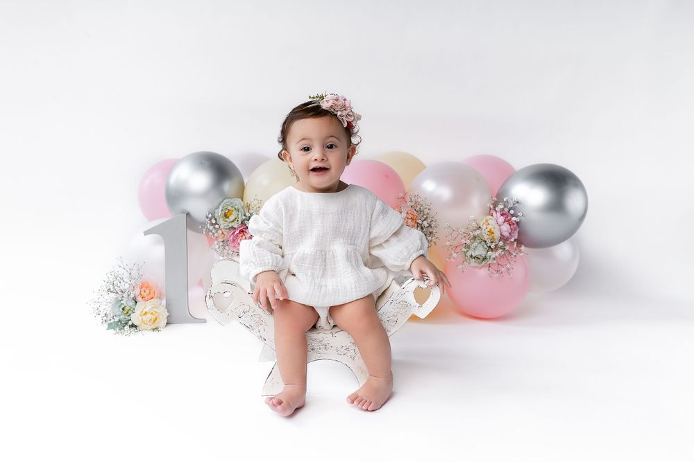Baby in white dress sitting among pink and silver balloons for a milestone photo shoot.