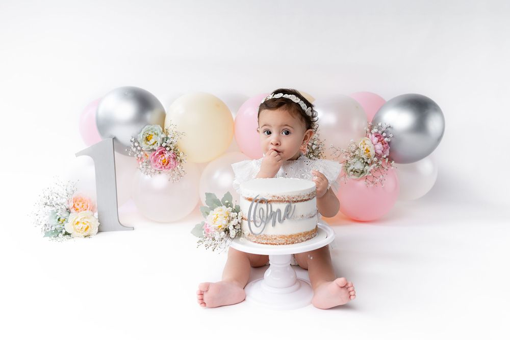 Baby in white dress poses with birthday cake and balloons for a cake smash photography session.