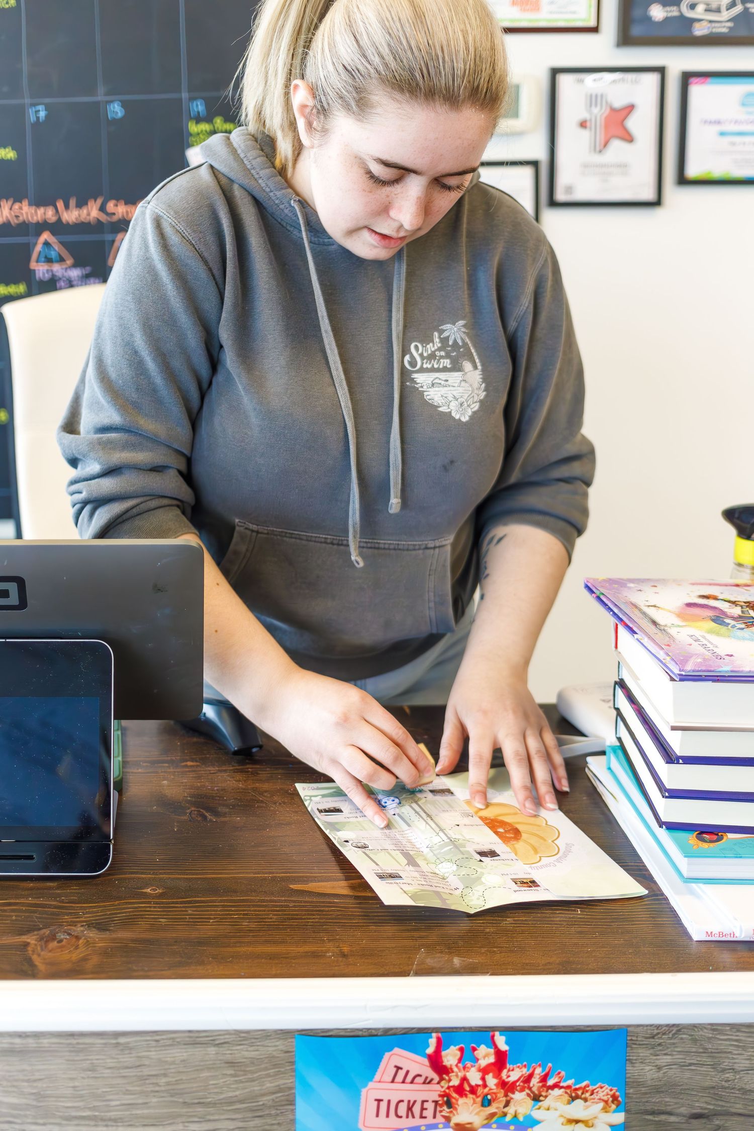 A retail worker processing items at a counter with books and a tablet display.