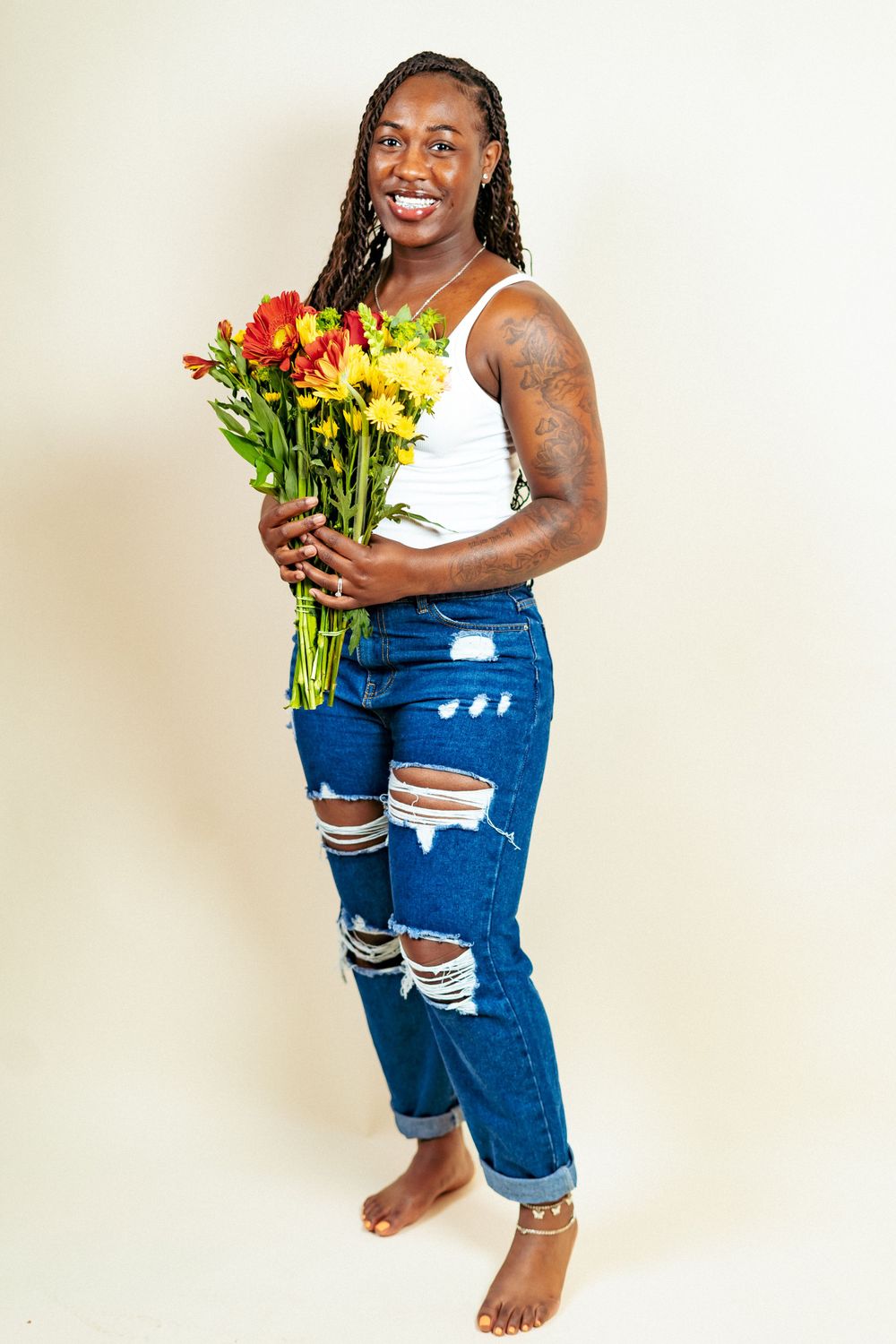 Person in distressed jeans and tank top holding colorful flowers against white background.