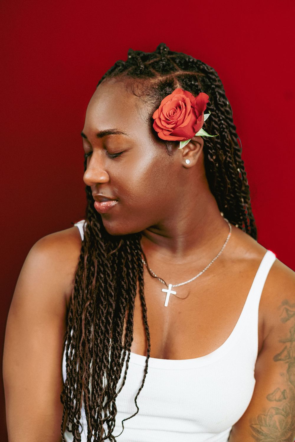 Person with braided hair wearing white top poses against red backdrop with coral rose accent.