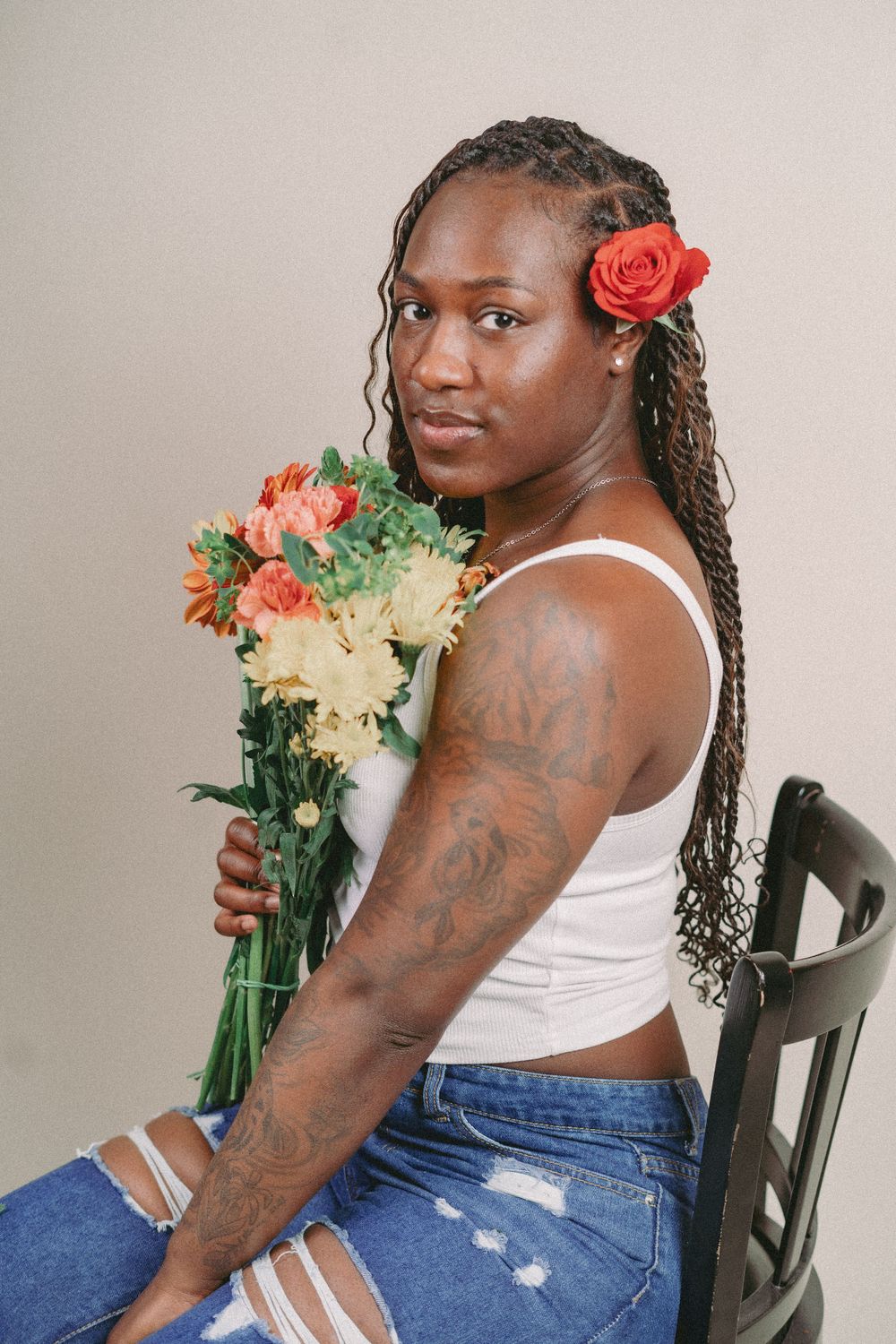 Person in white tank top and jeans poses with floral bouquet against light backdrop.