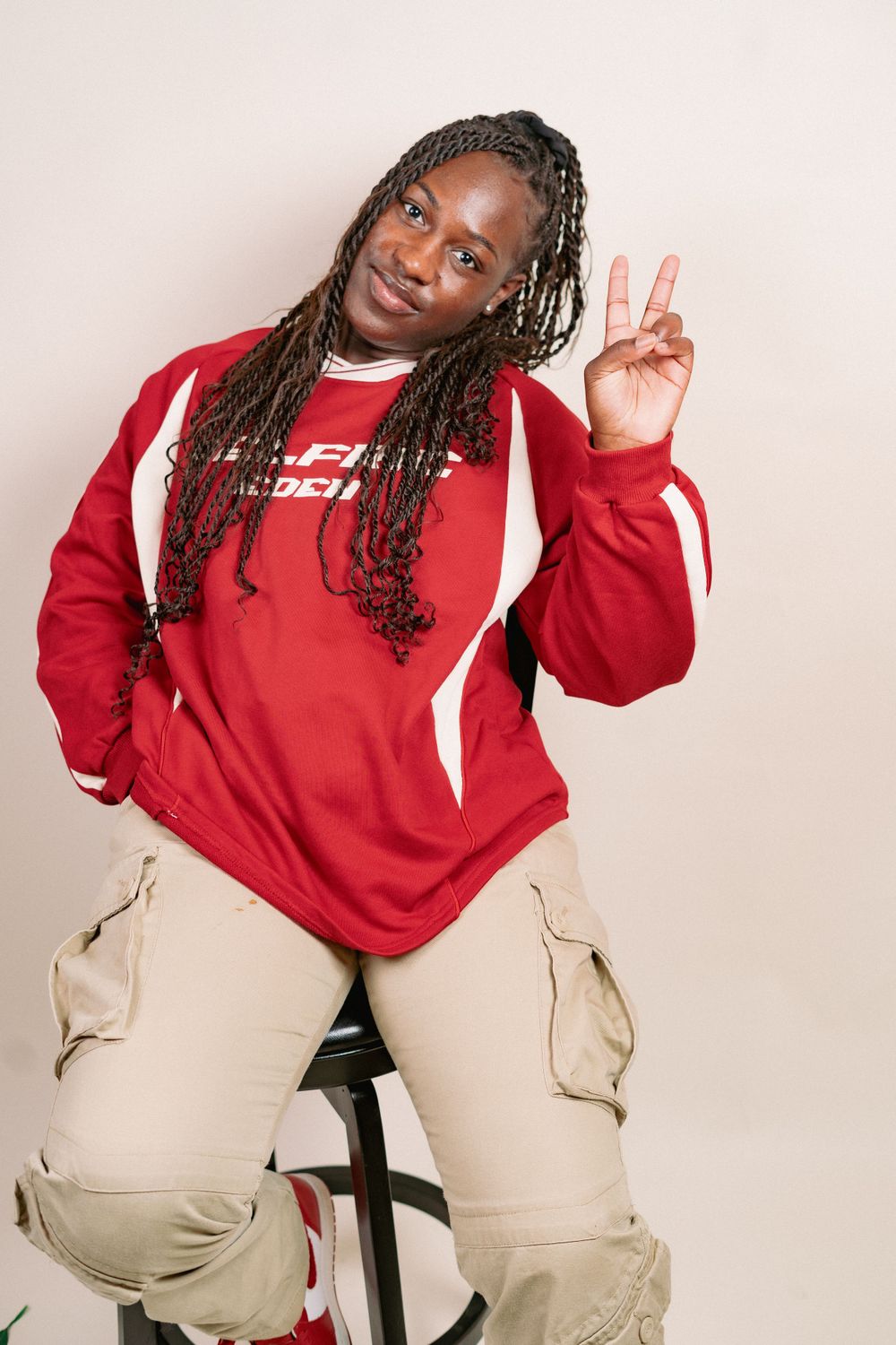Person in red sweater and cargo pants makes peace sign while seated on black chair.
