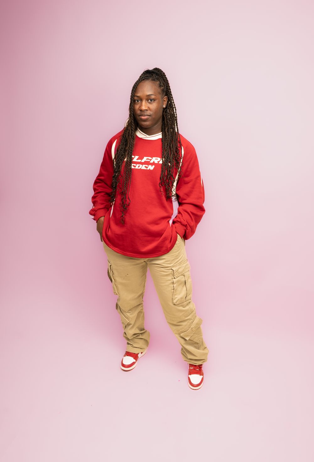 Model in red sweatshirt and khaki pants poses confidently against pink background.