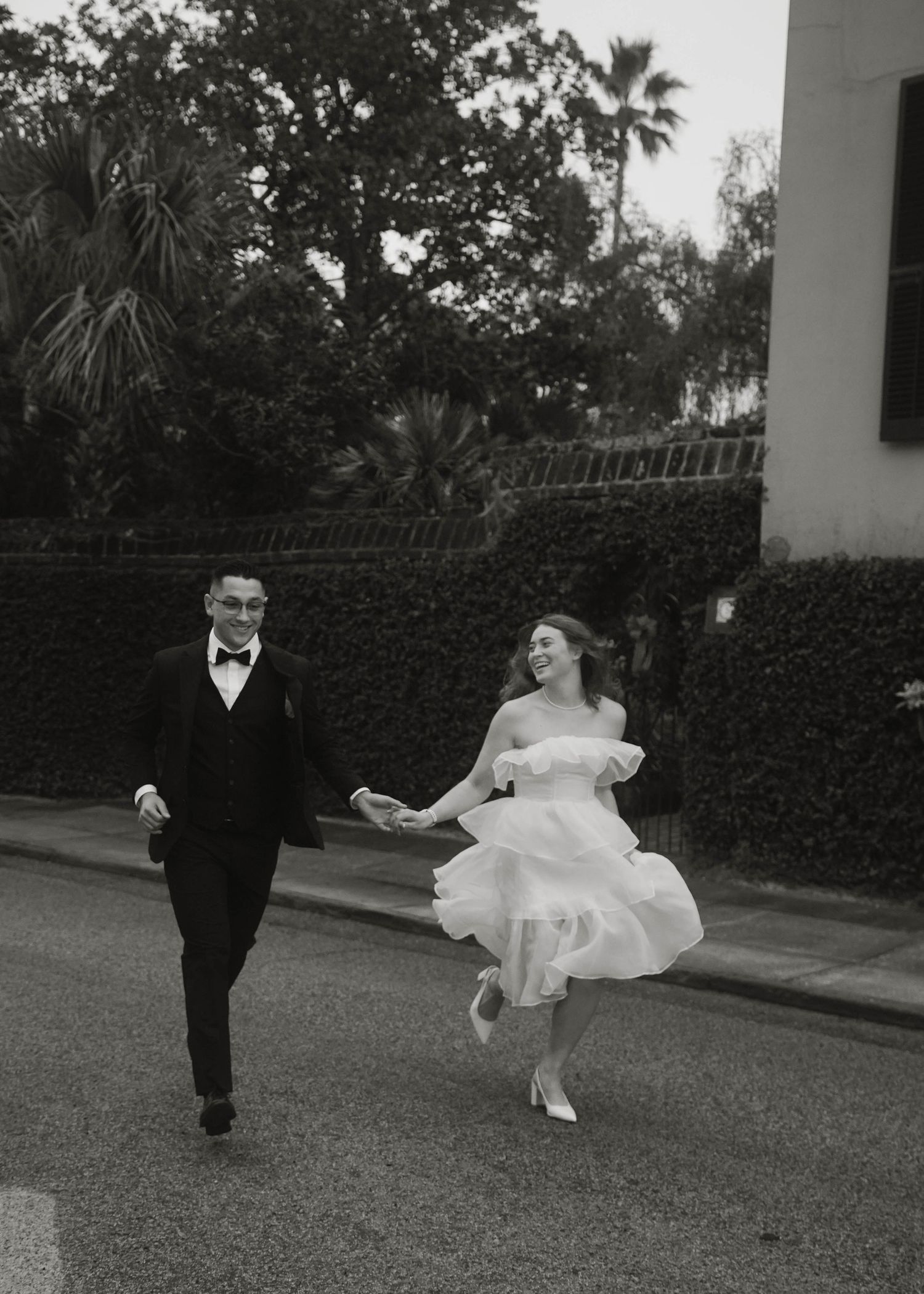 A couple in wedding attire playfully run down a palm tree-lined street in black and white.