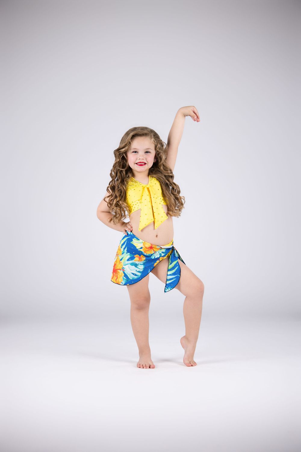 Young girl in yellow bikini top and blue floral sarong dancing barefoot against white background.