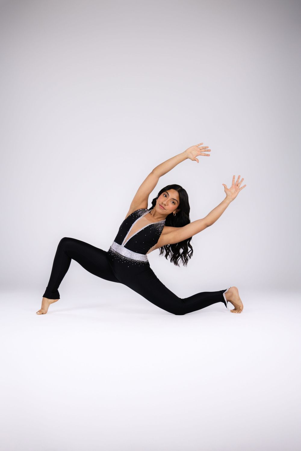 A dancer in a black jumpsuit performs a dramatic lunge pose with arms raised gracefully on a white background.