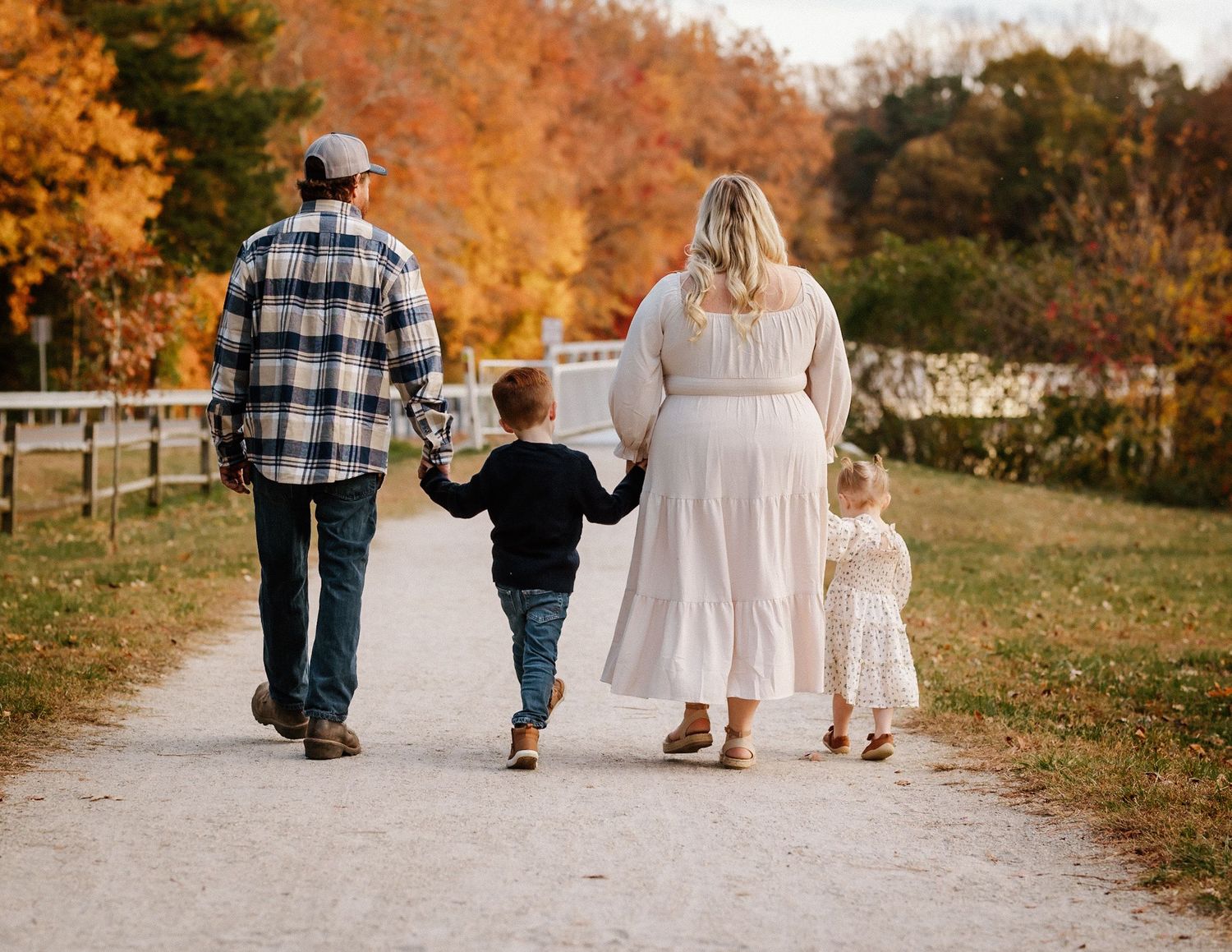 A family of four walks hand-in-hand down a scenic autumn path surrounded by colorful fall foliage.