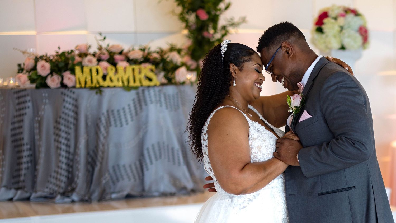 A couple shares an intimate dance moment at their wedding reception with floral decorations and 'Mr & Mrs' sign in background.