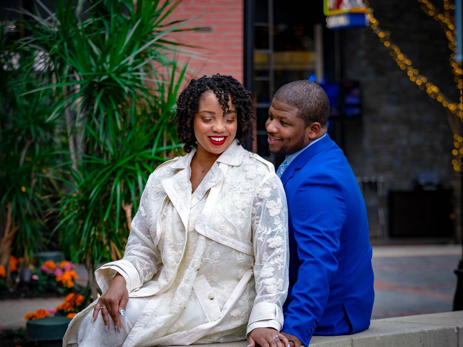 A happy couple in white and blue attire pose together outdoors near foliage and decorative lighting.