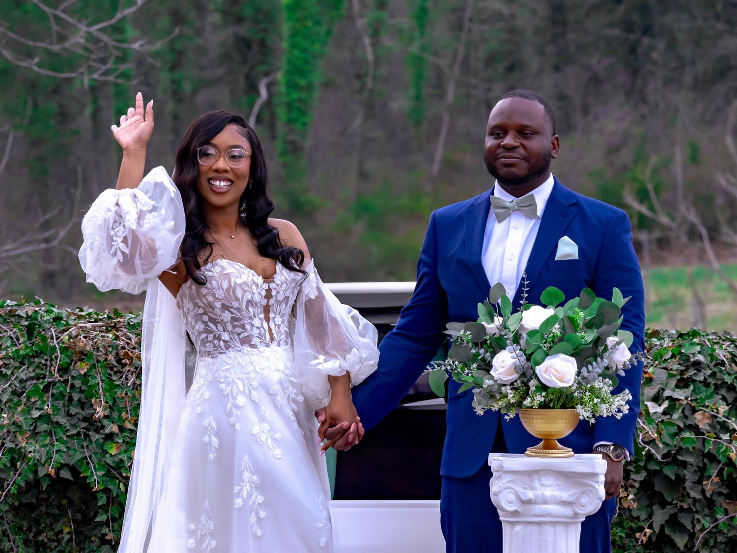 Elegant bride waves while holding hands with groom in navy suit outdoors in a garden setting with greenery.