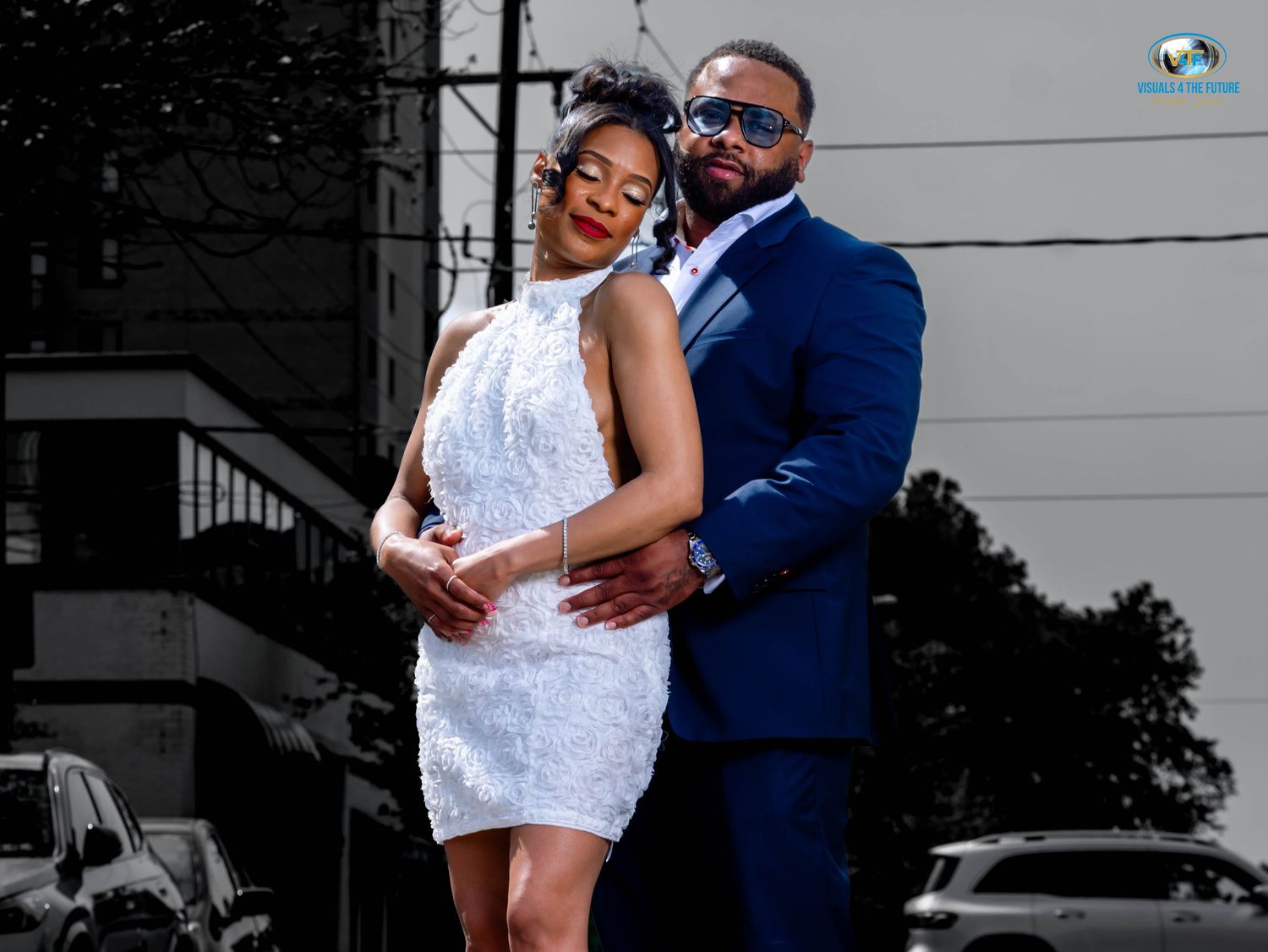 A couple poses together outdoors, with one wearing a white lace dress and the other in a navy blue suit.