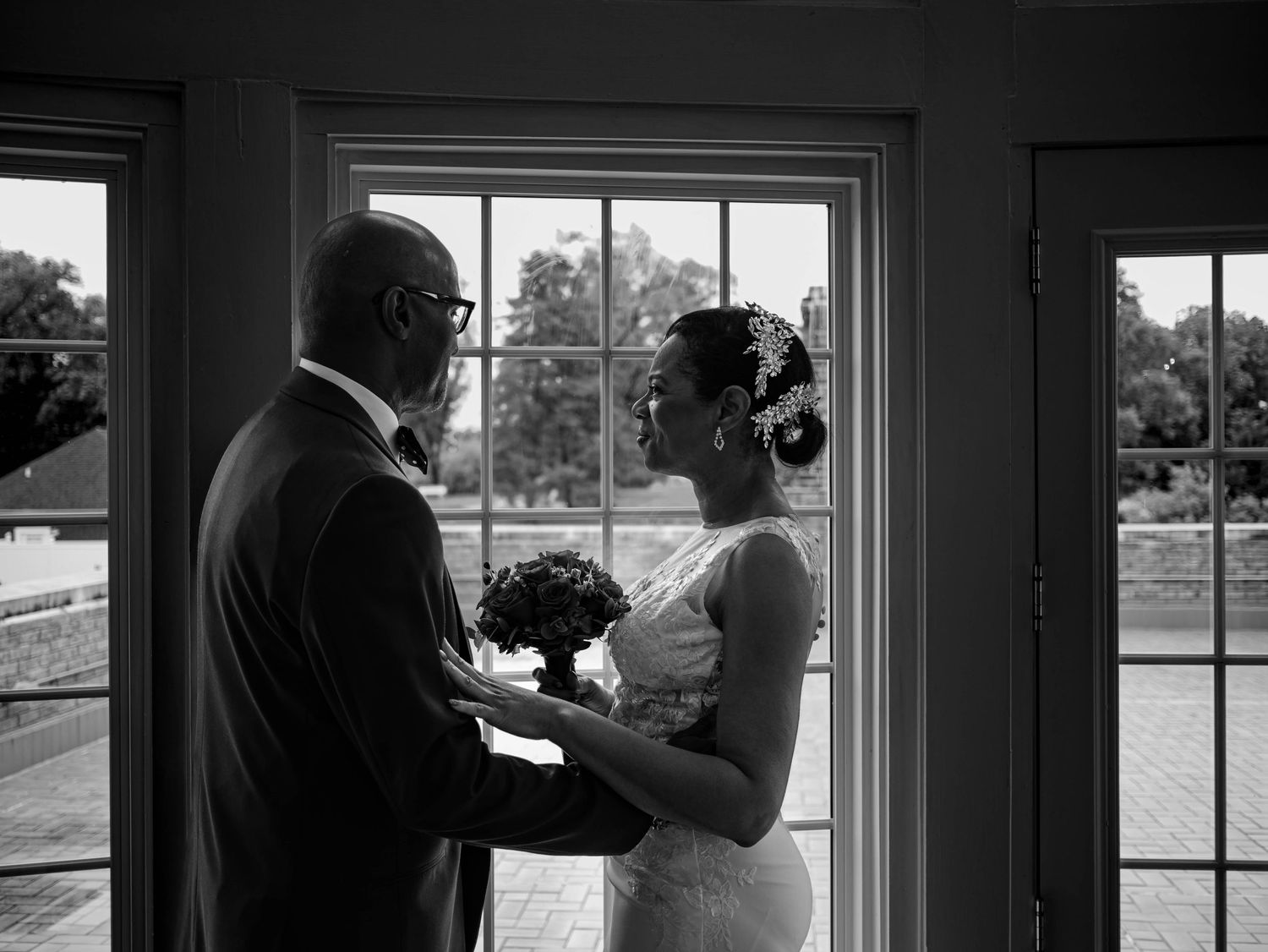 A tender black and white wedding moment captured near a window as two people share an intimate exchange with flowers.