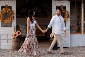 Playful couple in summer outfits holding hands and dancing by a rustic white building.