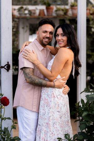 Couple in coordinated outfits embraces near white garden trellis with red roses.