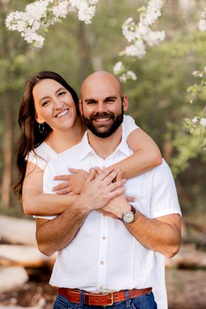 Couple in white outfits embracing outdoors with flowering trees in background.
