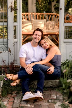 A couple poses together on rustic stone steps in front of a vintage greenhouse.