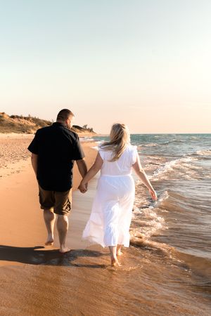 Couple walks hand in hand along shoreline during golden hour beach photo session.
