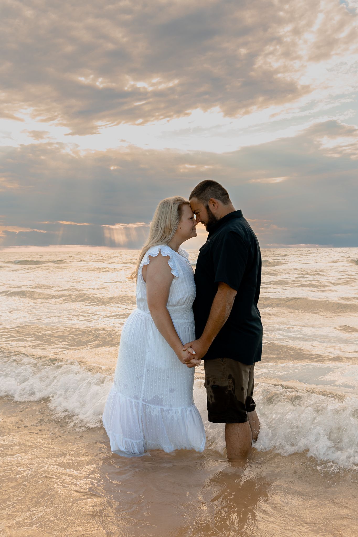 Couple poses in crashing waves during dramatic beach sunset photoshoot.