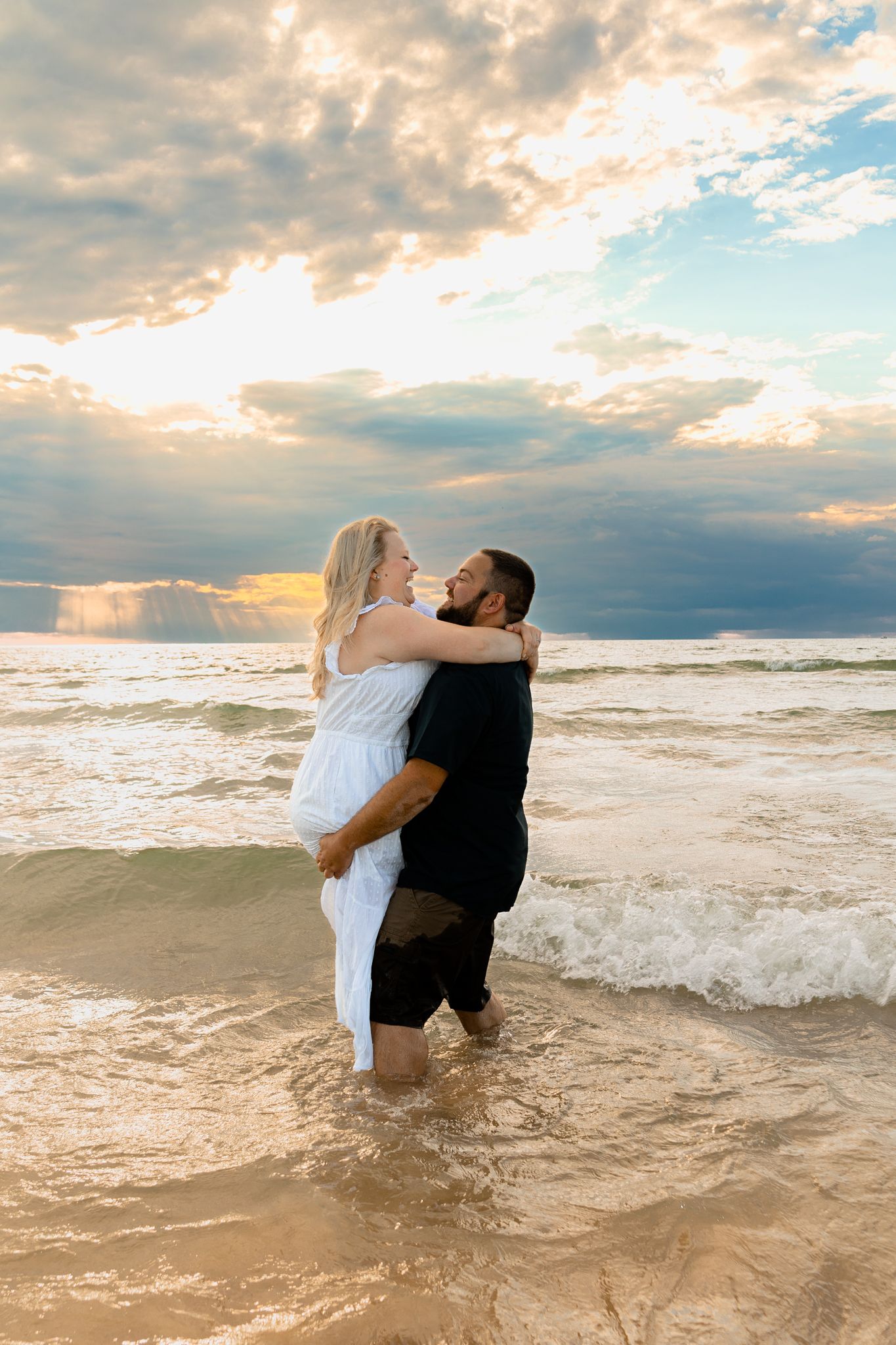 Romantic couple embraces while standing in ocean waves during sunset photo session.