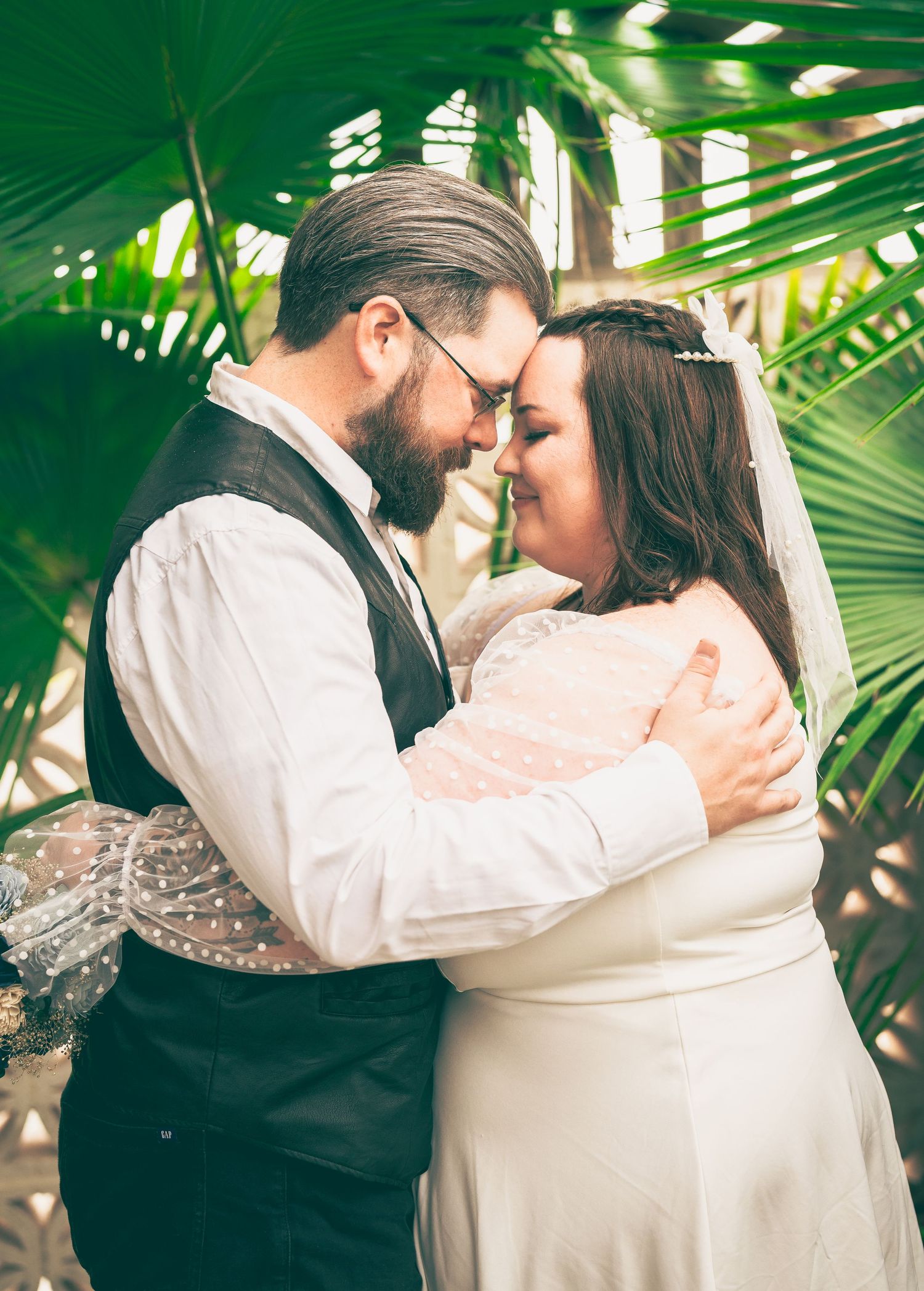 Romantic wedding couple embraces under tropical palm fronds in a greenhouse setting.