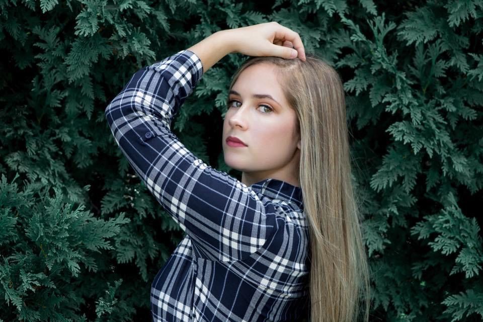 A young woman with long blonde hair poses against a lush green hedge in a plaid shirt.