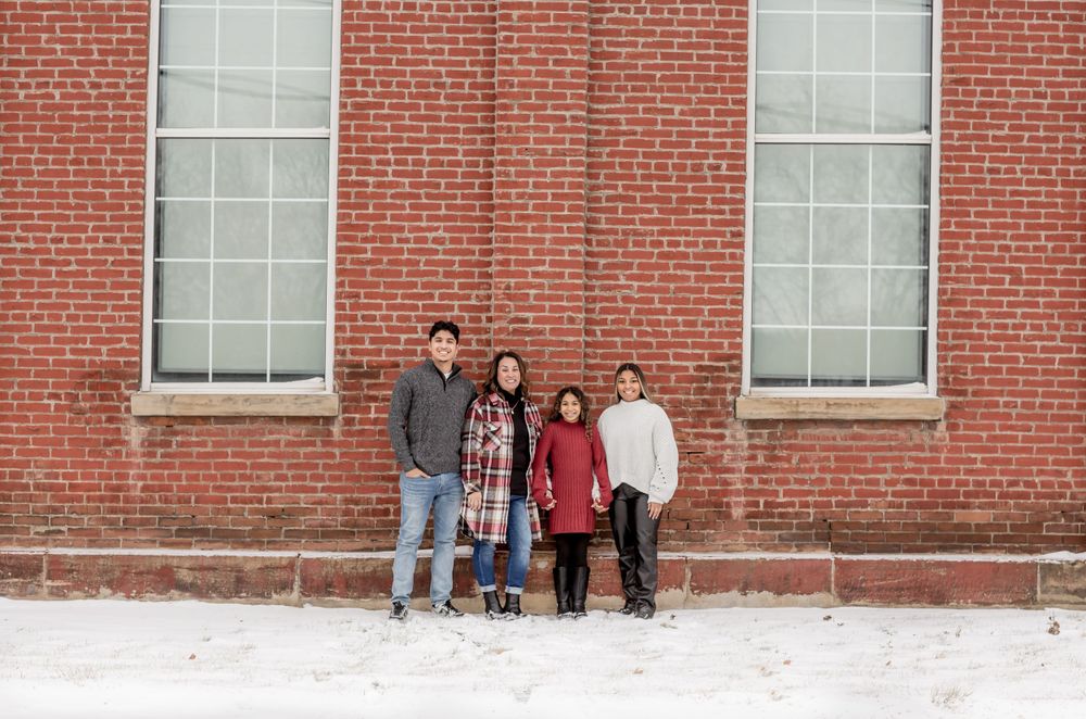 A family of four stands together in front of a large red brick building in winter snow.