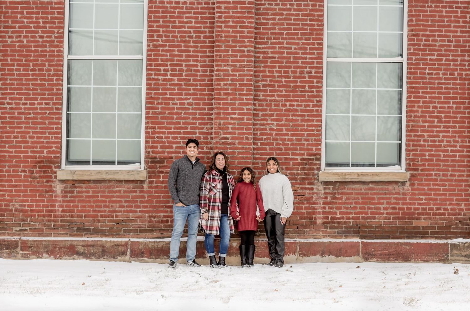 A family of four stands together in front of a large red brick building in winter snow.