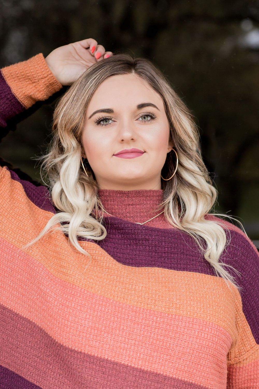 A young woman in a colorful striped sweater poses outdoors with her hand in her wavy blonde hair.