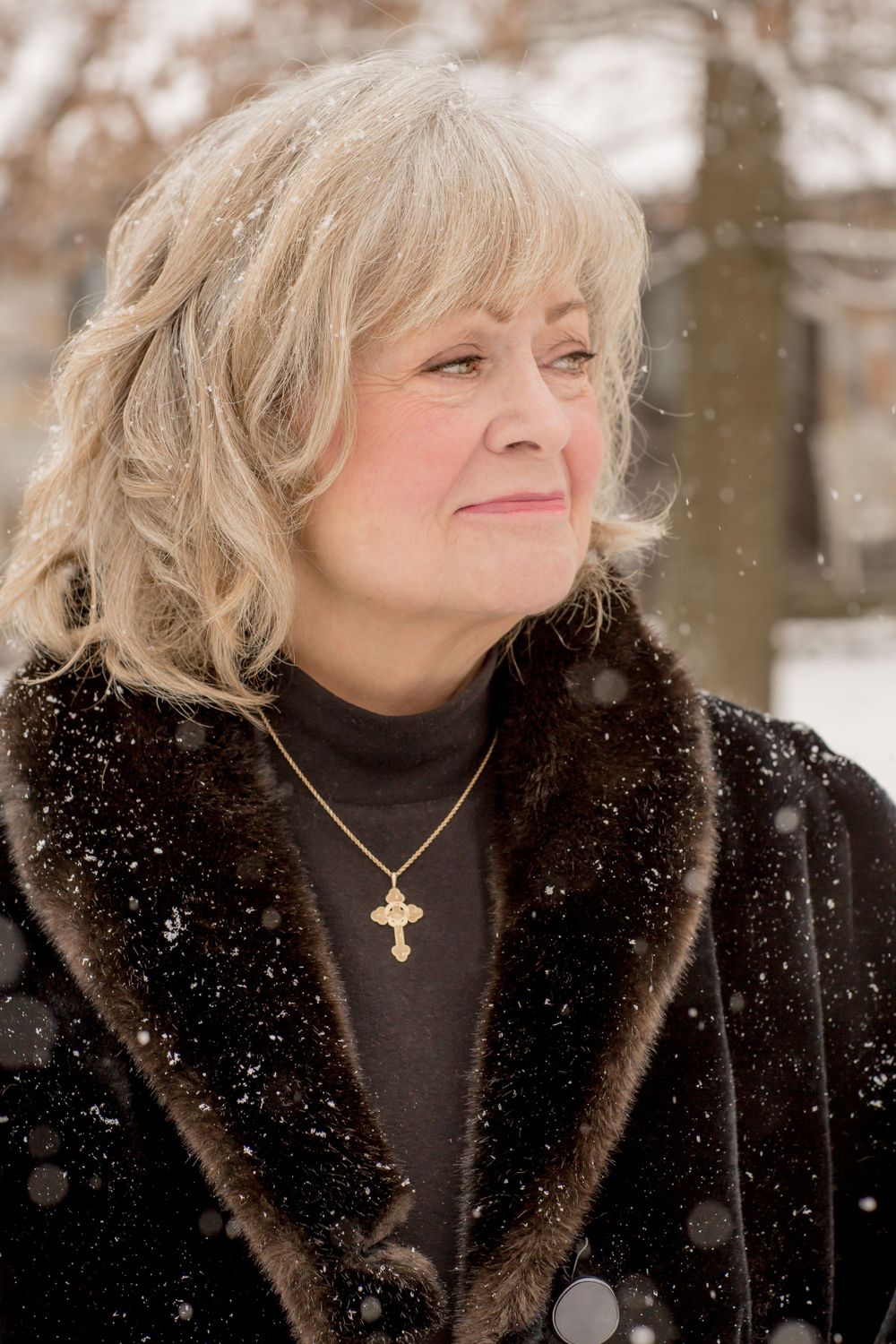 An elegant older woman in a dark fur coat wearing a gold cross necklace stands in falling snow.