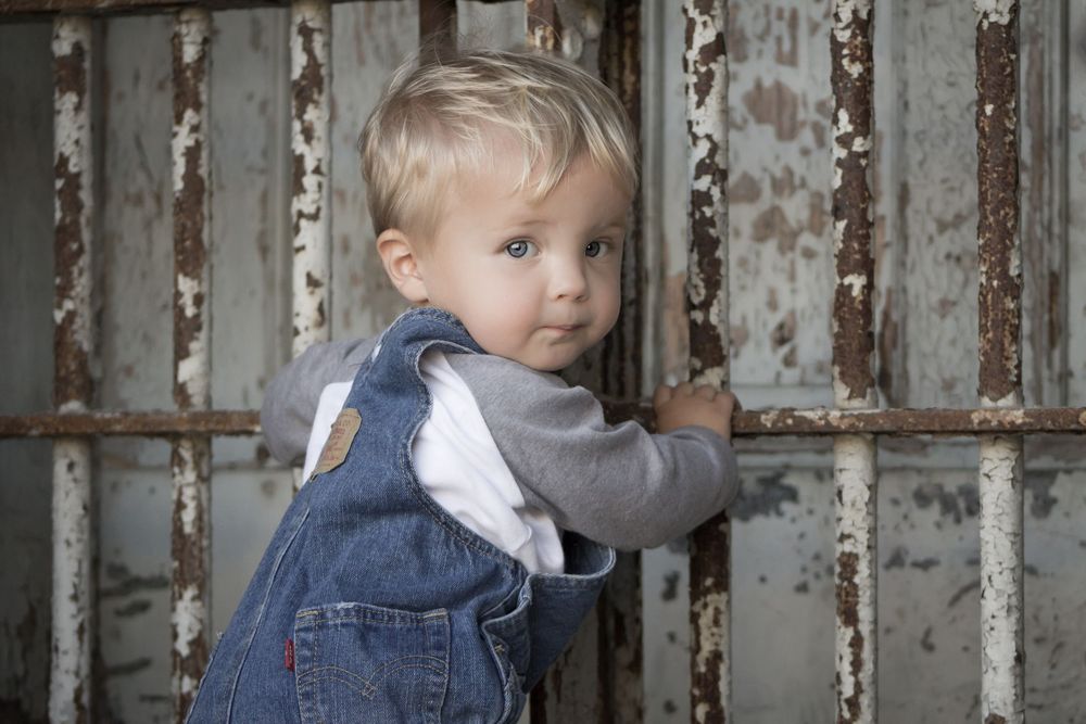 A blond toddler boy in denim overalls leans against a rustic peeling metal gate.