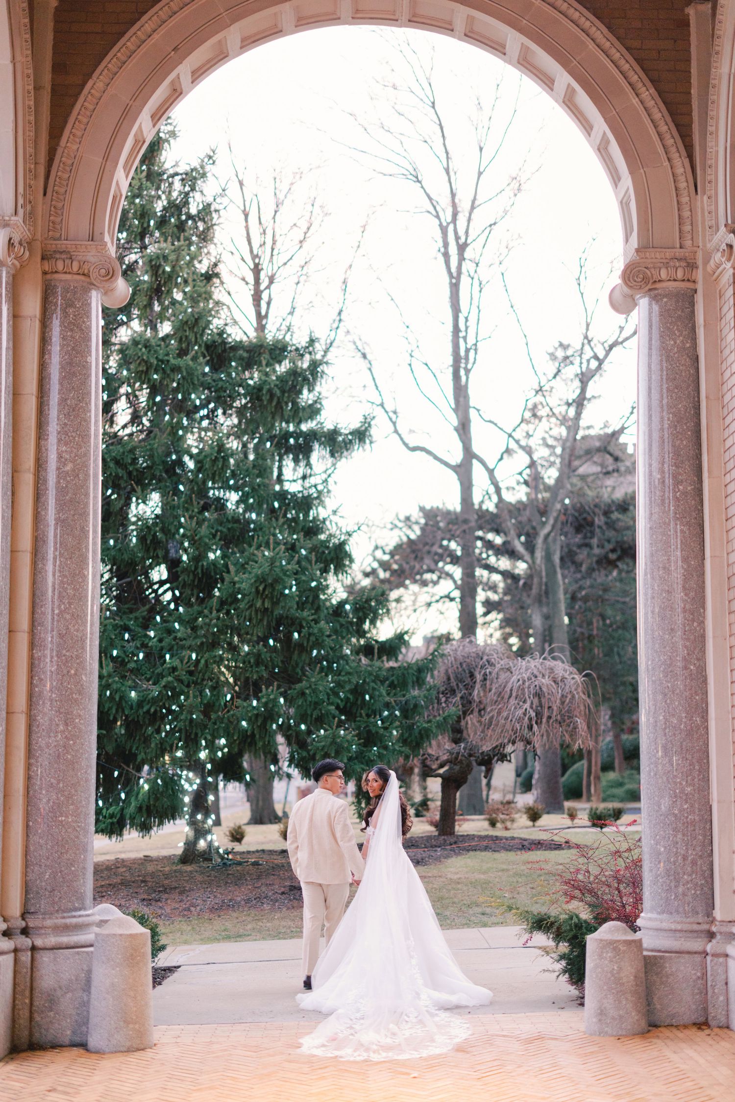 Wedding couple holding hands under ornate stone arch surrounded by lush garden trees and greenery.