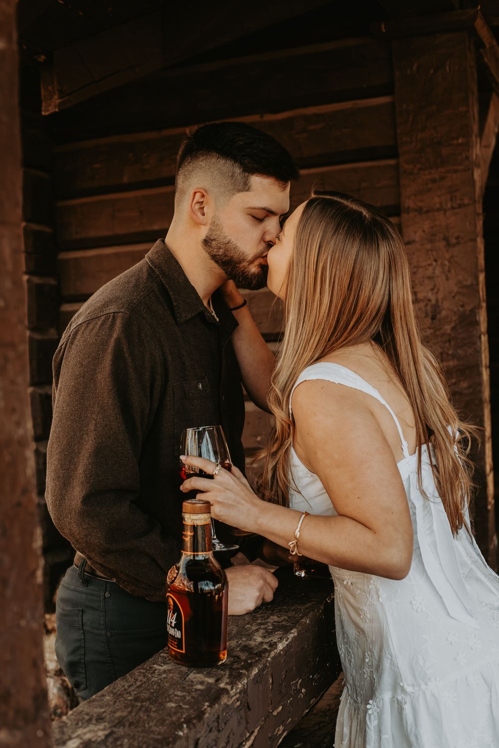 Intimate couple moment sharing kiss with wine glass and whiskey bottle in rustic setting.