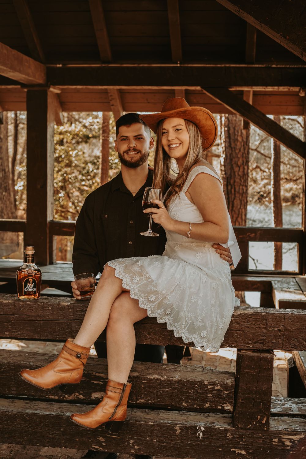 Couple enjoying drinks and laughter in rustic cabin setting with cowboy boots and white dress.