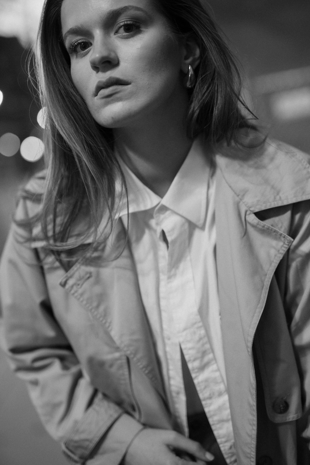 Black and white portrait of someone in a classic trench coat and white collared shirt looking away pensively.