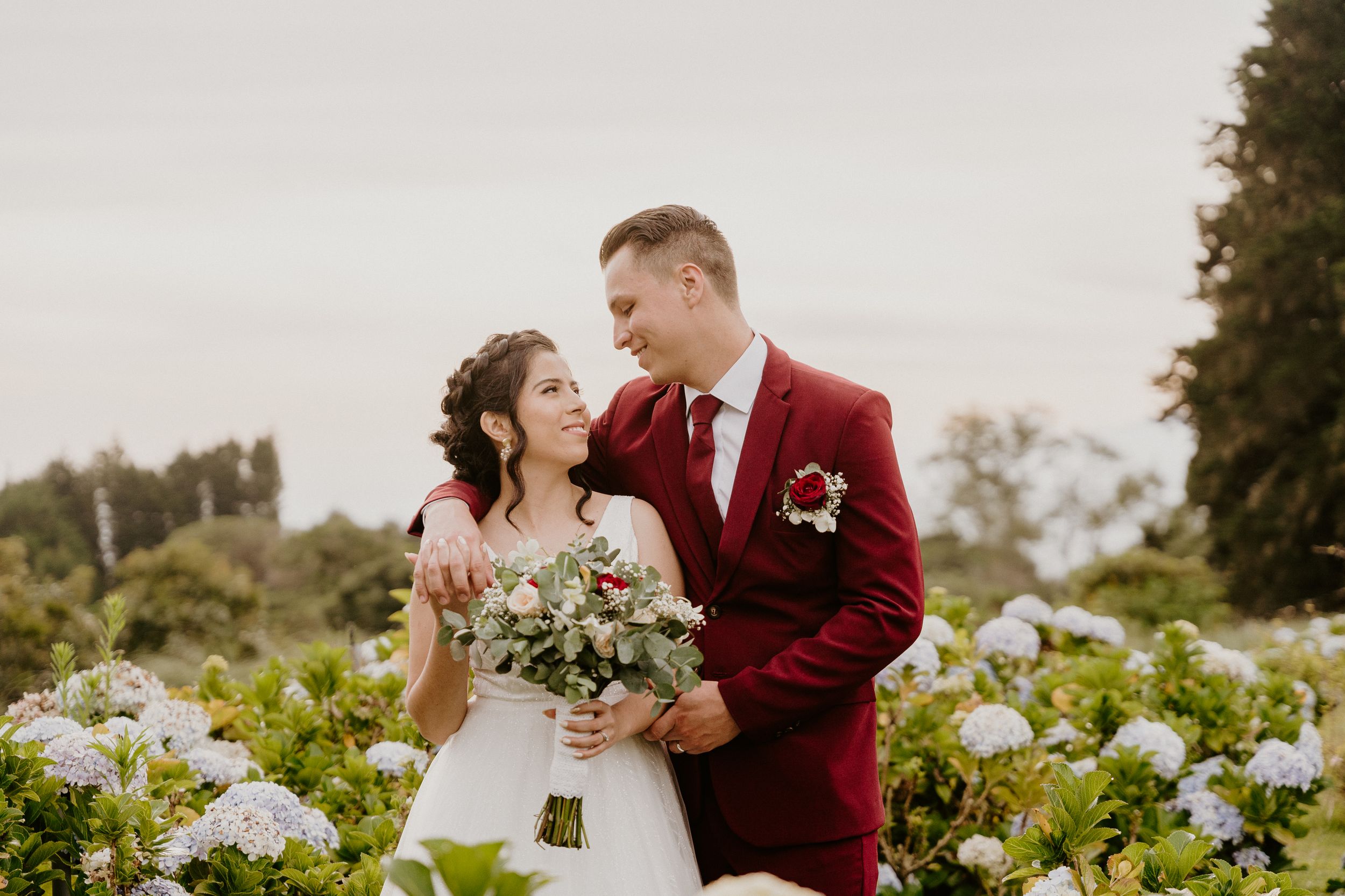 A wedding couple poses in a garden with white hydrangeas, the groom wearing a striking burgundy suit. Heredia, Boda, la yoyita, sesión fotos