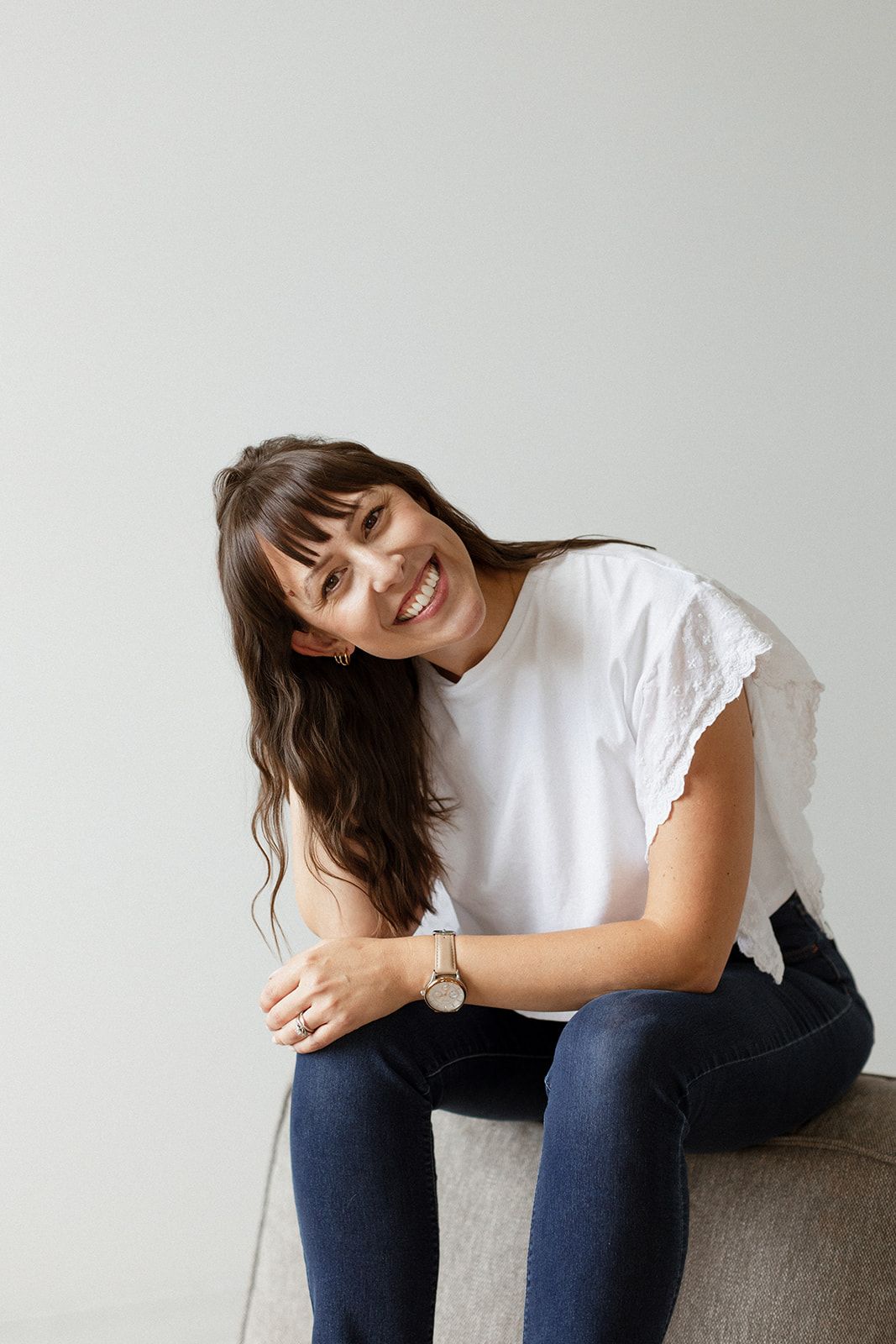 Person wearing casual white t-shirt and jeans sits on bench with joyful expression against light background.