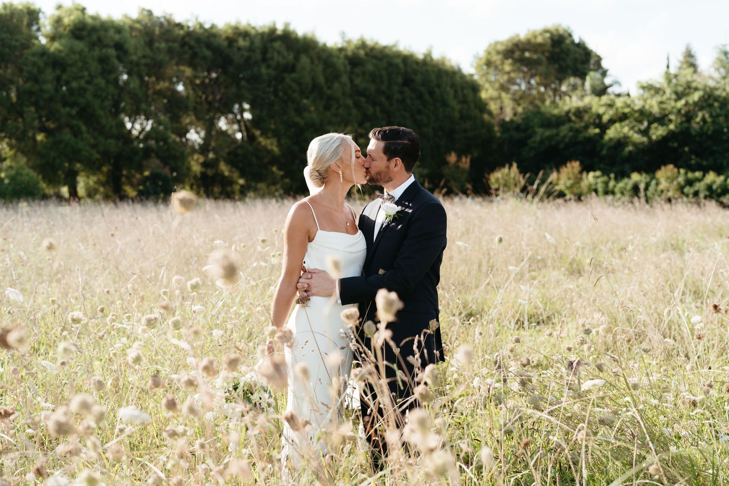A couple shares a tender moment in a sunlit wildflower meadow on their wedding day at Markovina Vineyard Estate in Auckland.