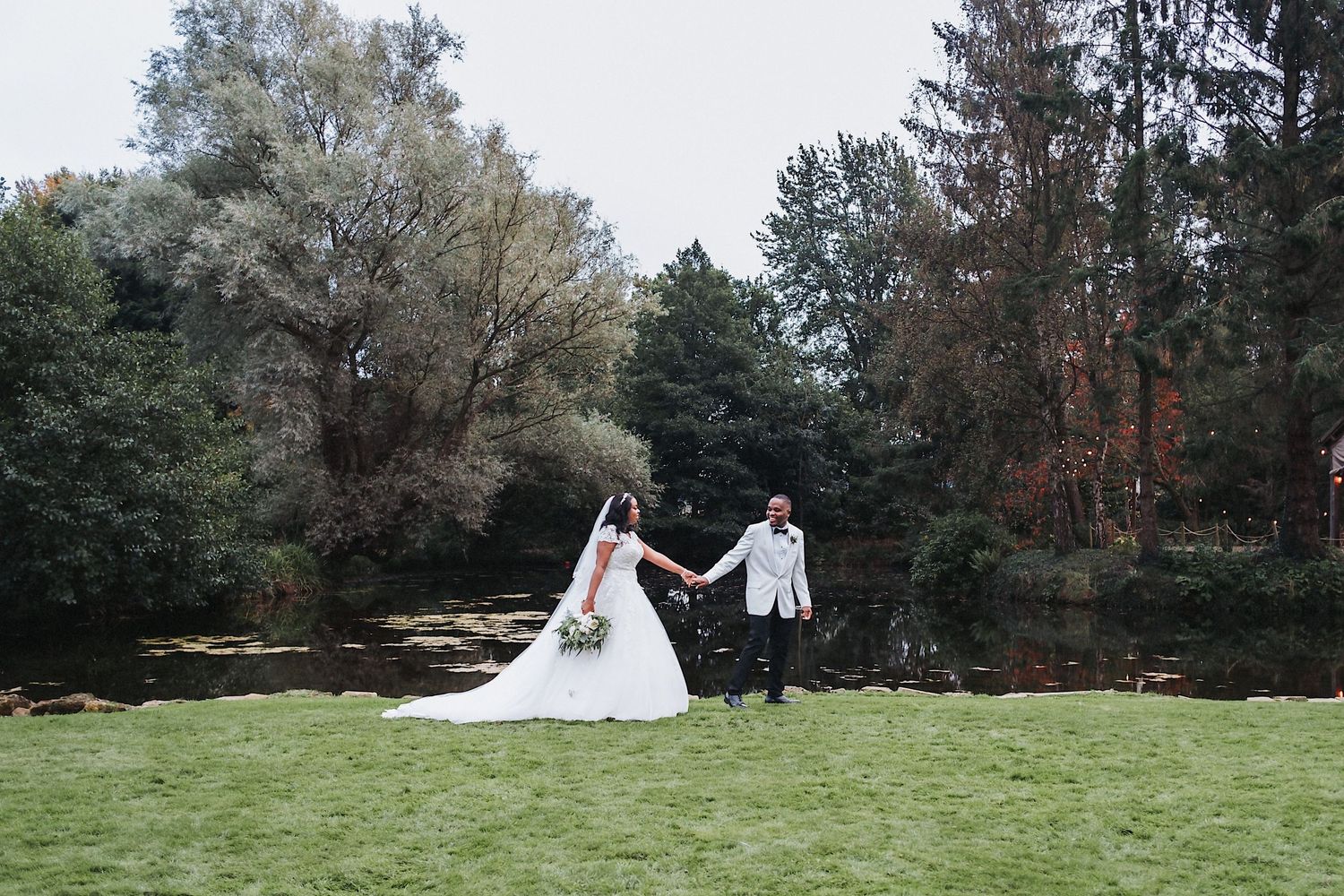 A wedding couple holds hands while walking across a grassy lawn surrounded by trees.