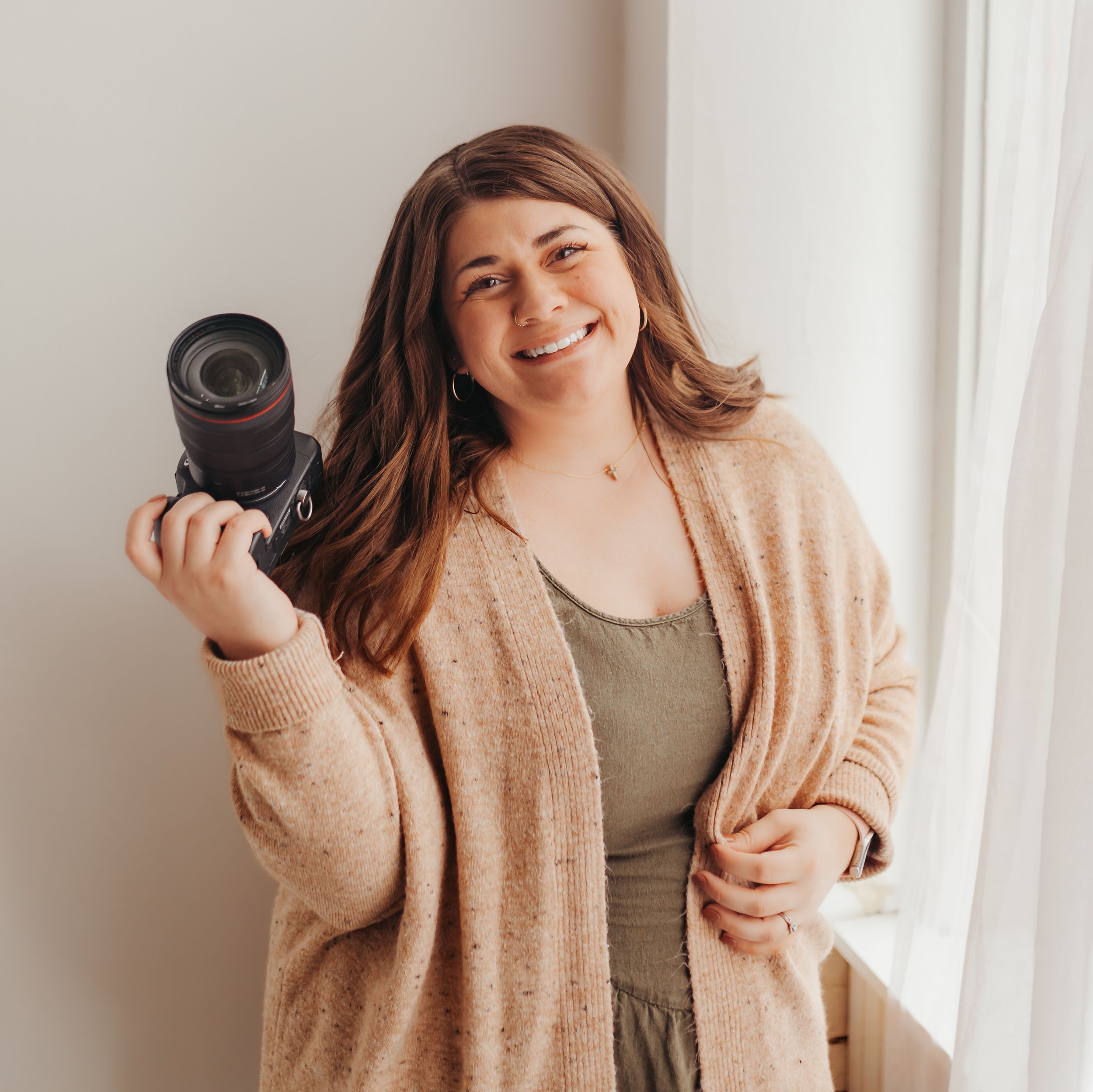 A photographer in a white sweater and hat sits casually on a stool holding a camera against a rust-colored wall.