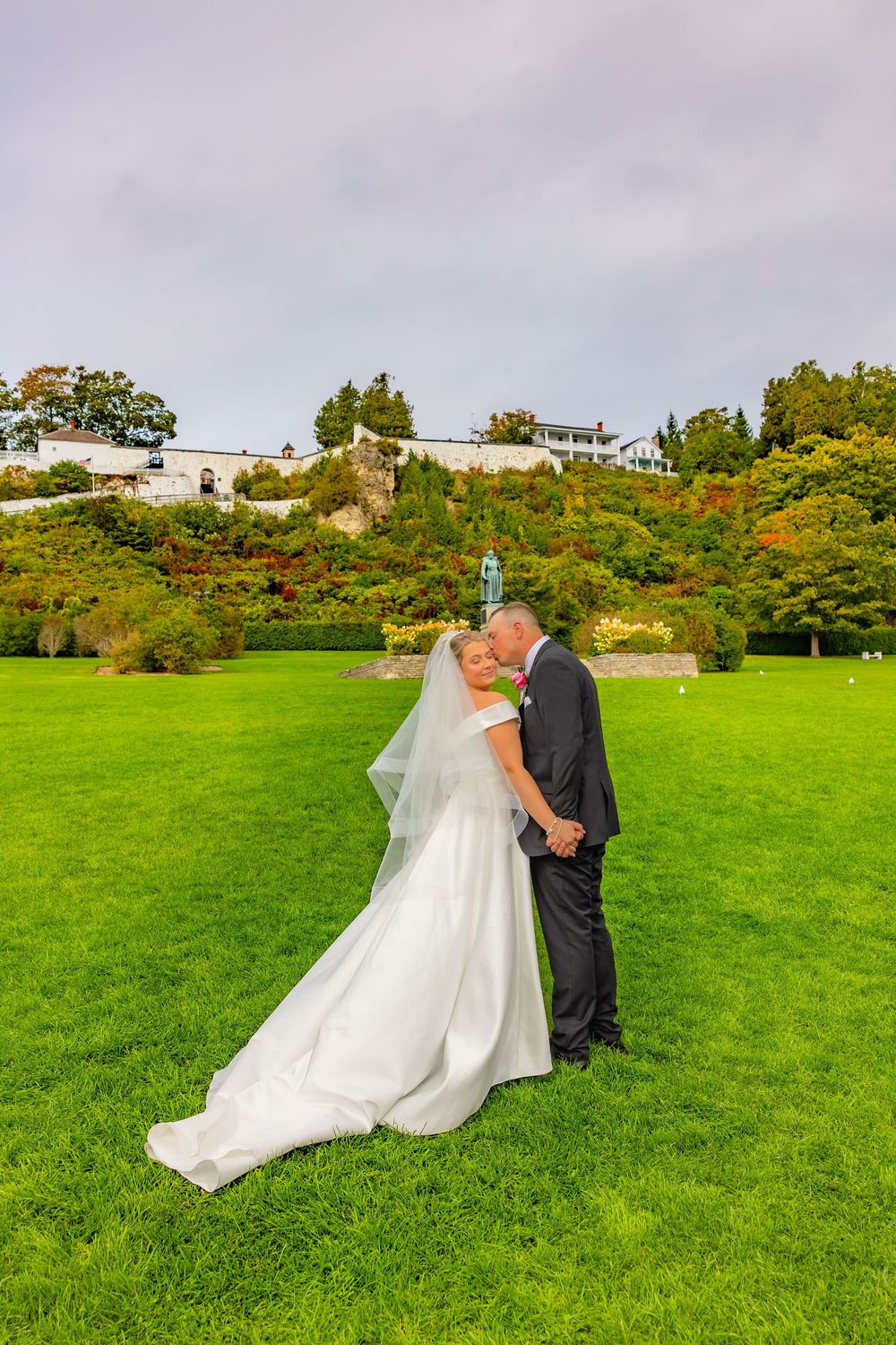 A couple shares a romantic moment in their wedding attire on a lush green lawn with trees in the background.