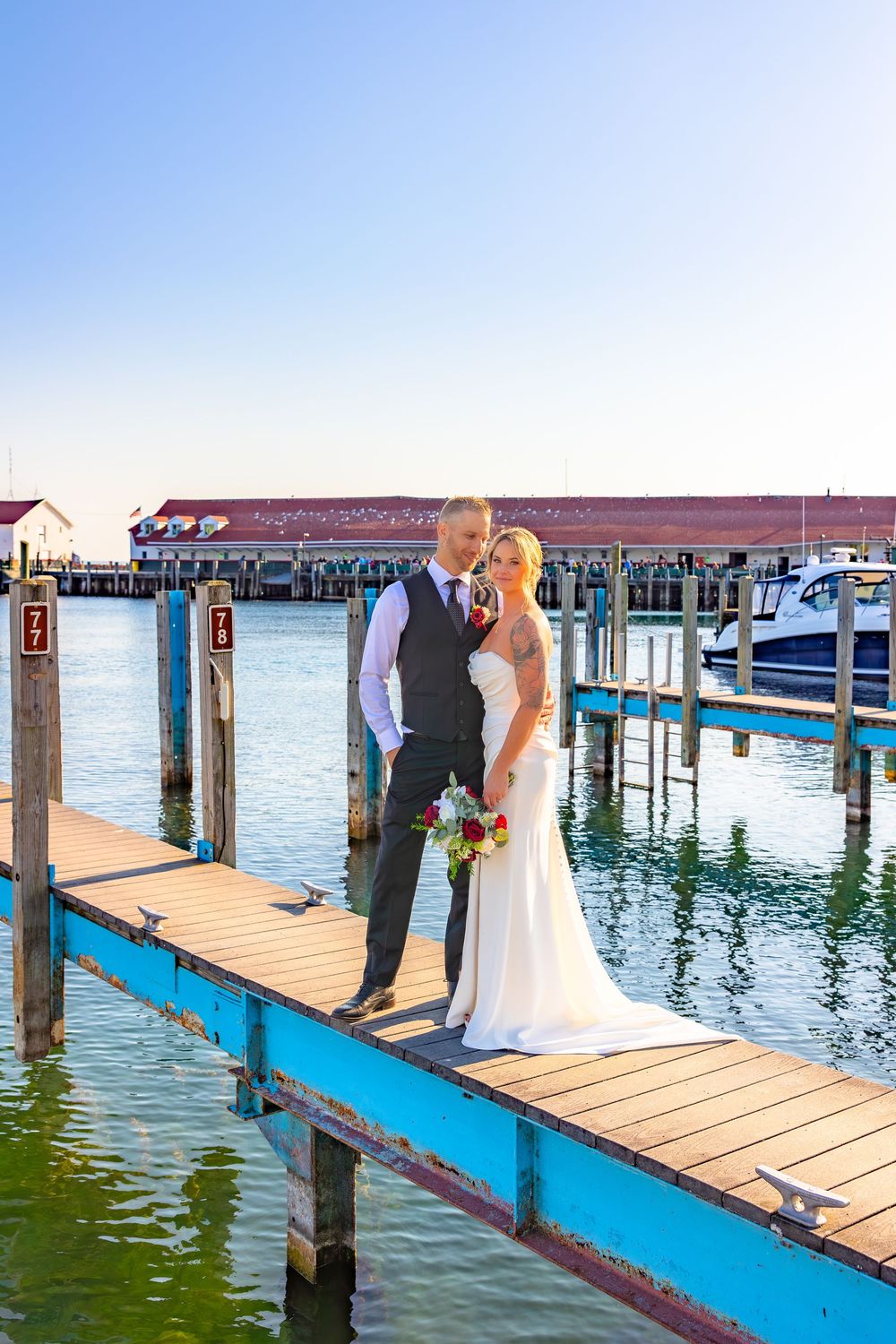 Wedding couple poses on a wooden dock overlooking blue water with a red-roofed building in the background.