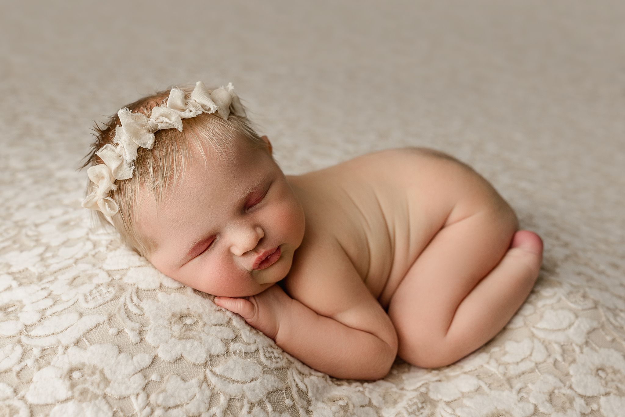 A sleeping newborn baby lies peacefully on soft white lace fabric in a professional studio portrait setting.