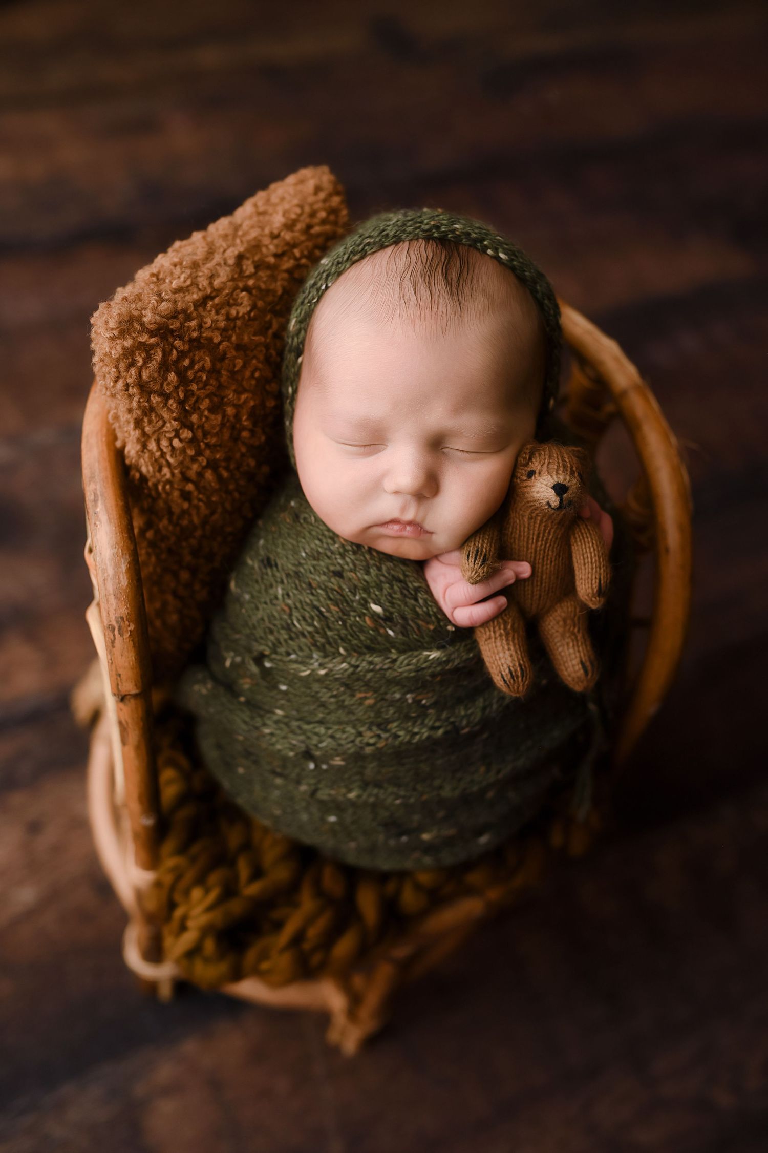Newborn baby wrapped in green knit swaddle holding tiny bear in wooden basket prop.