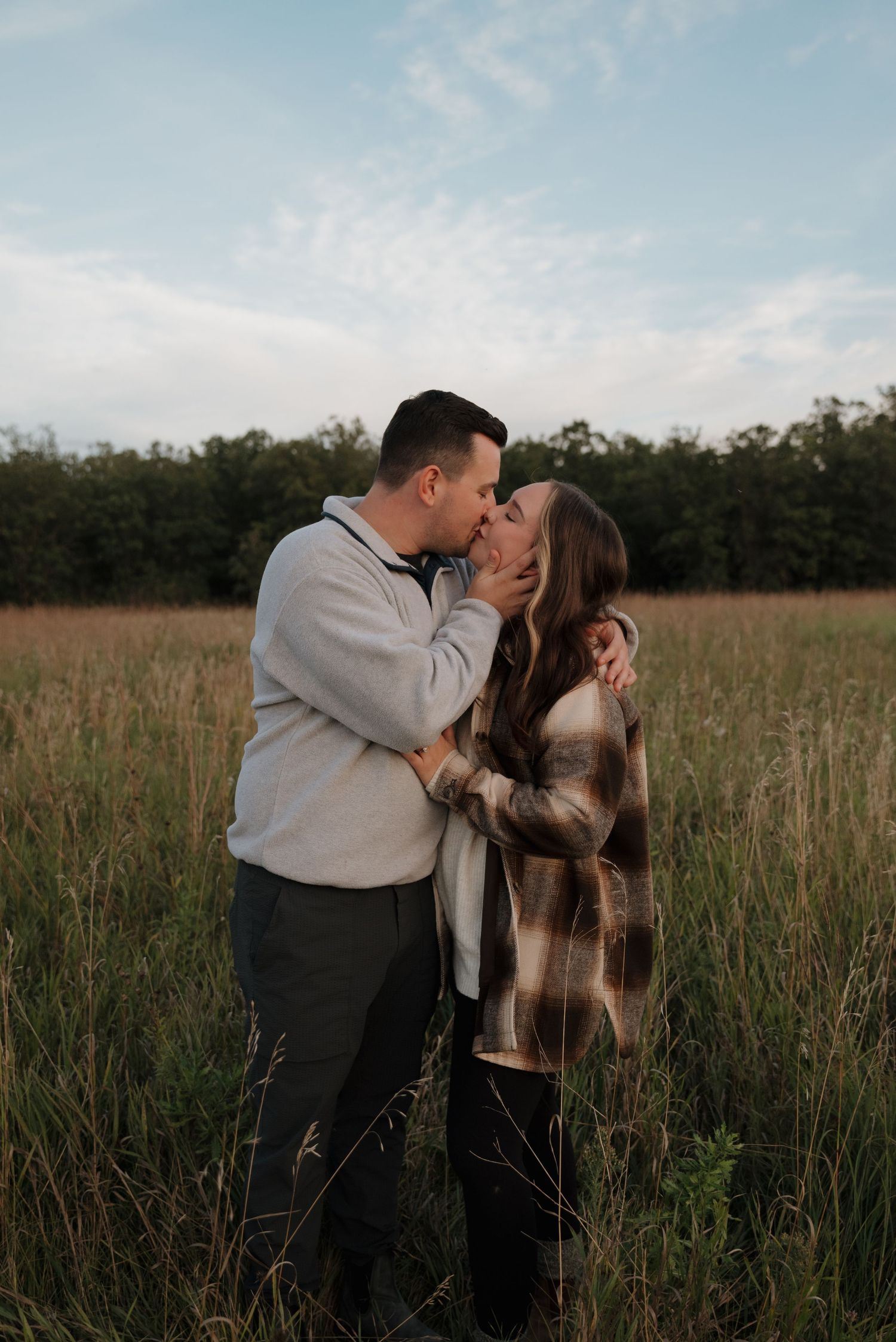 A couple shares romantic kisses in a tall grass field at sunset, creating intimate outdoor engagement photos.