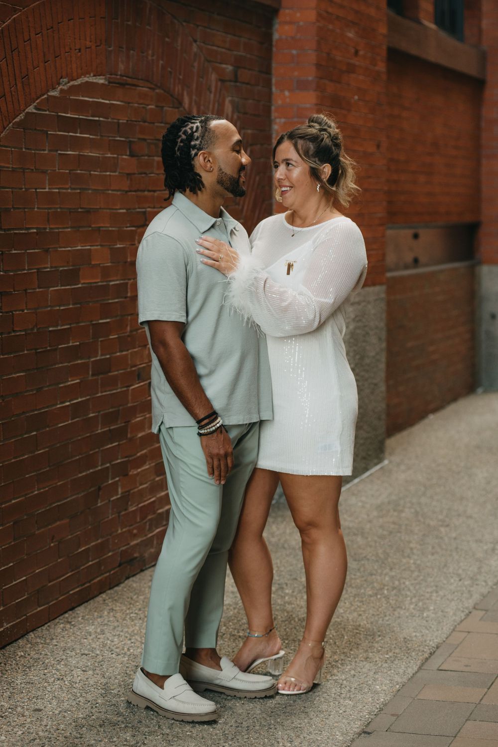 Couple laughing and embracing against a red brick wall, man in sage green and woman in white.