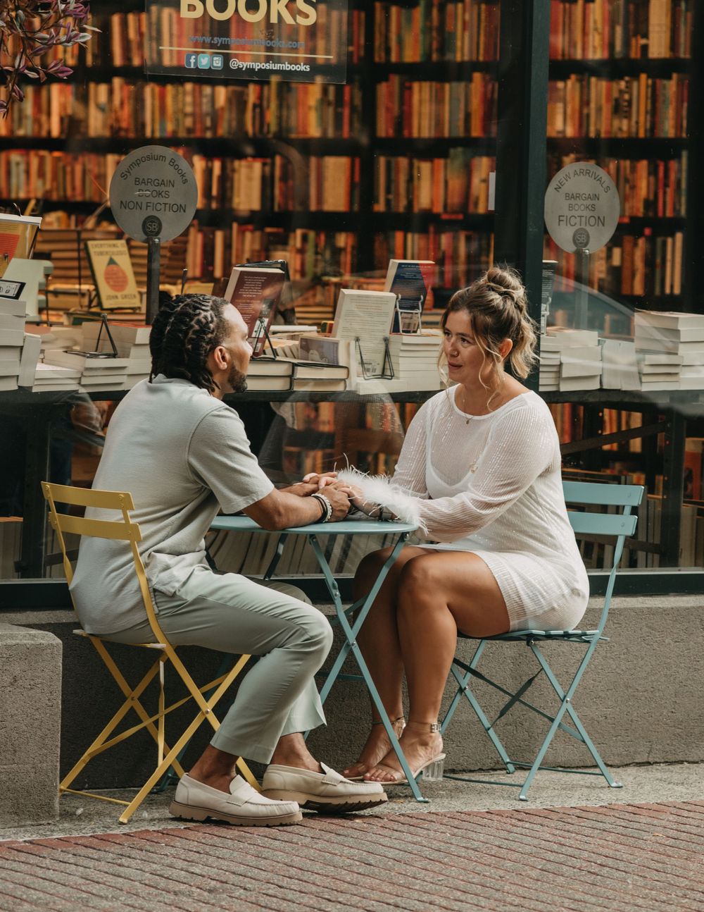 Couple holding hands at a bistro table outside a bookstore with colorful book displays.